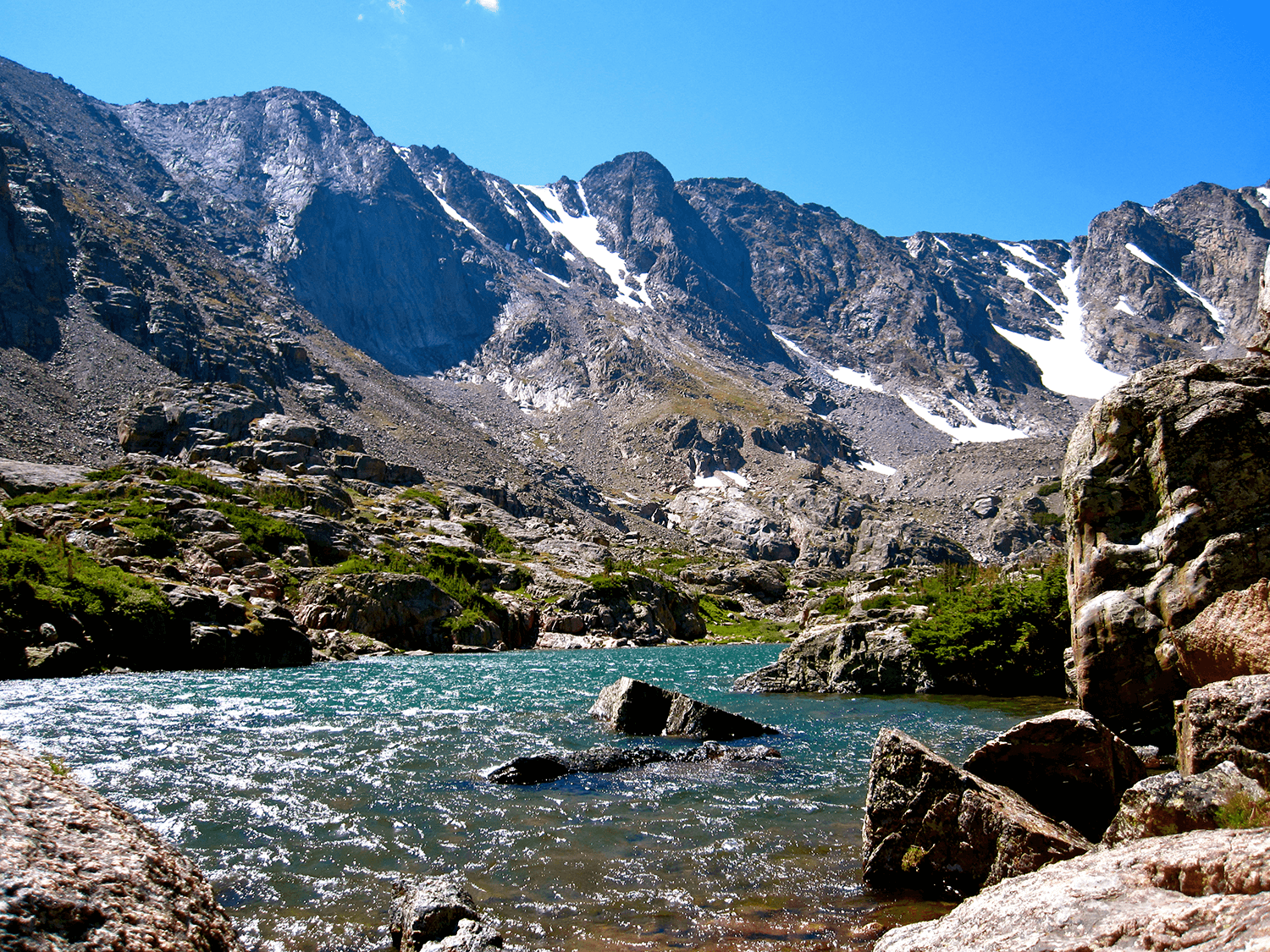 Mountain landscape with snow patches, rocky slopes, and a clear blue lake in the foreground.