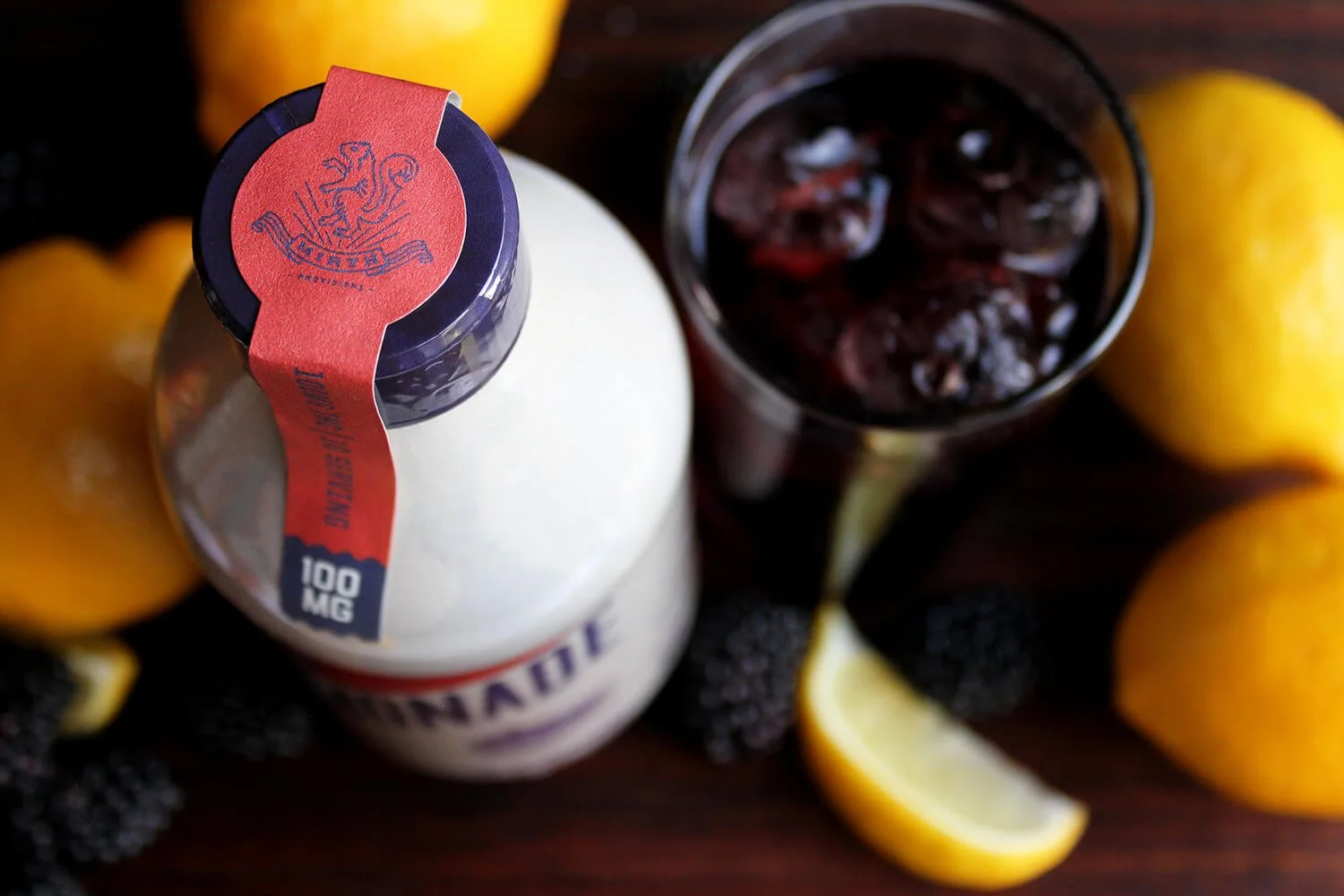A bottle of CBD oil surrounded by blackberries, lemon slices, and whole lemons, with a glass of berry juice in the background on a dark wooden surface.
