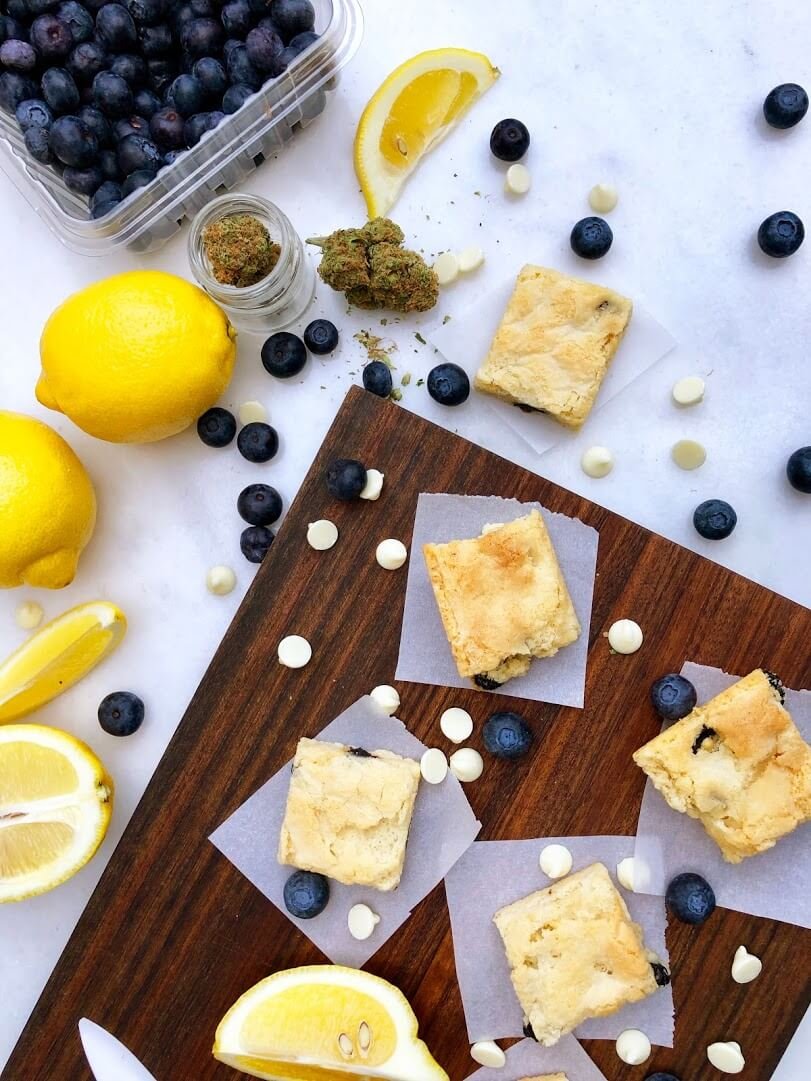 Lemon blueberry scones on parchment paper on a wooden board, with fresh blueberries, lemon slices, and a small jar of cannabis buds nearby.