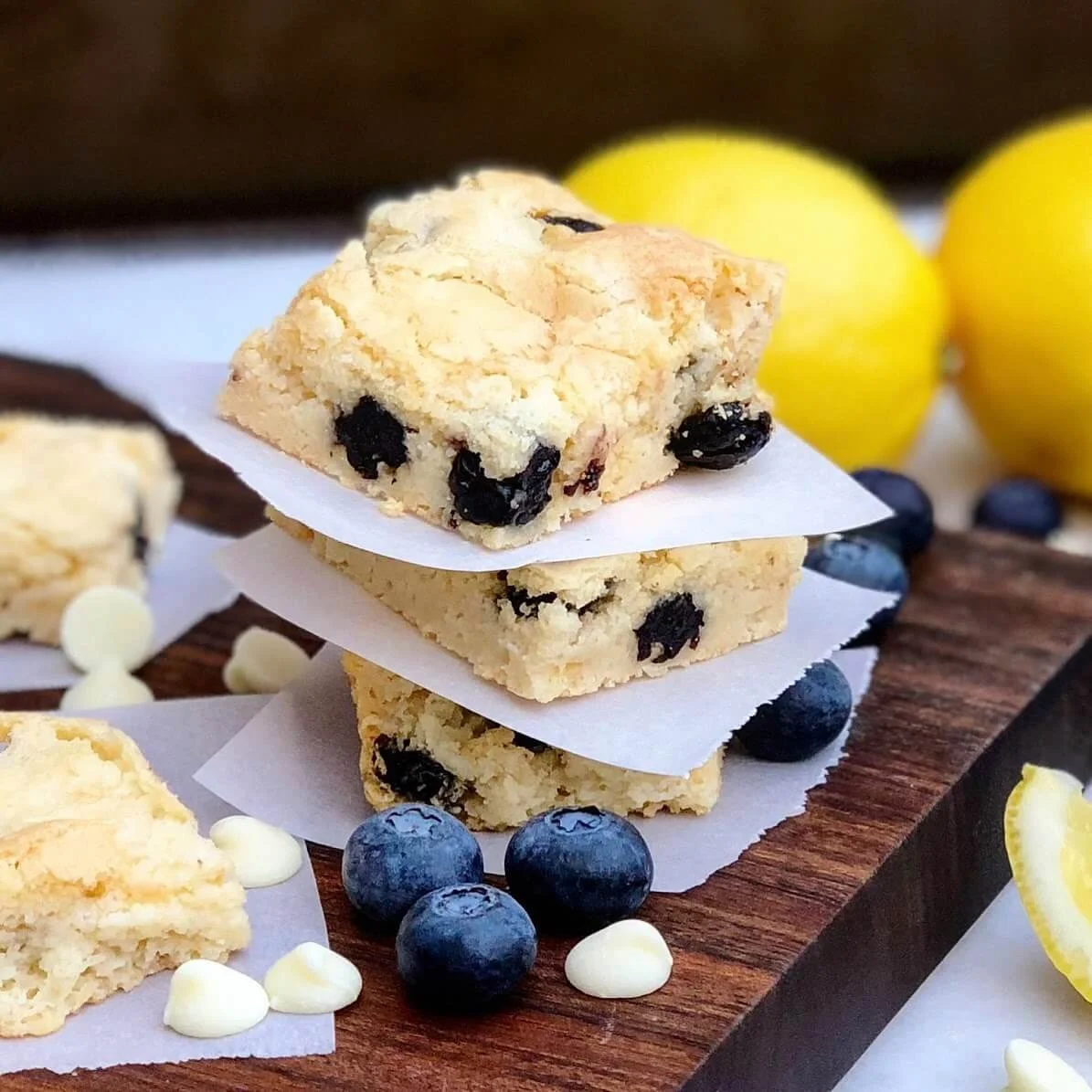 Three blueberry crumb bars stacked on parchment paper on a wooden serving board, with fresh blueberries and lemon slices in the background.