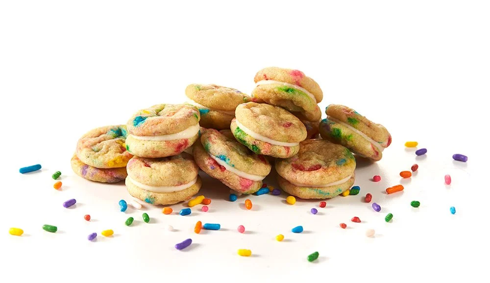A pile of colorful cookies with white frosting and rainbow sprinkles on a white background.