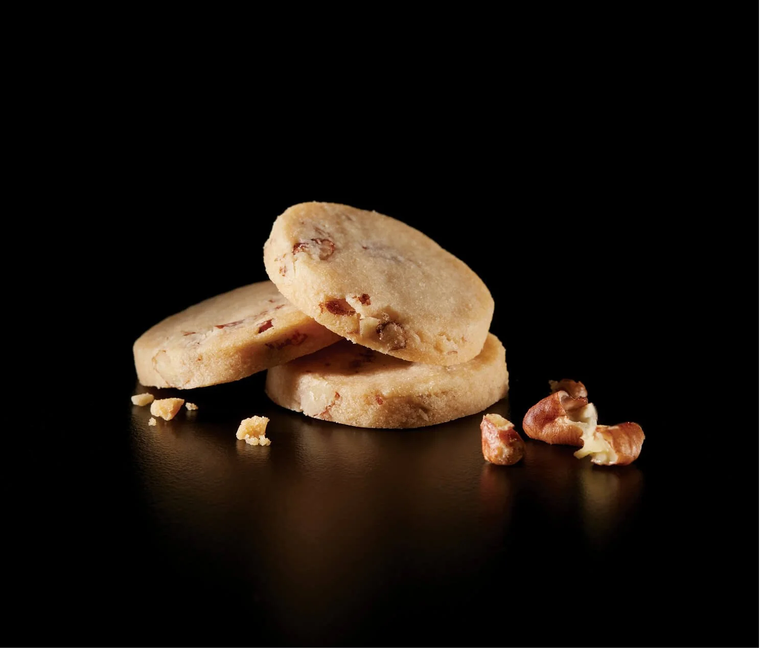 Three chocolate chip cookies stacked on a black surface with scattered chopped nuts around them, against a black background.