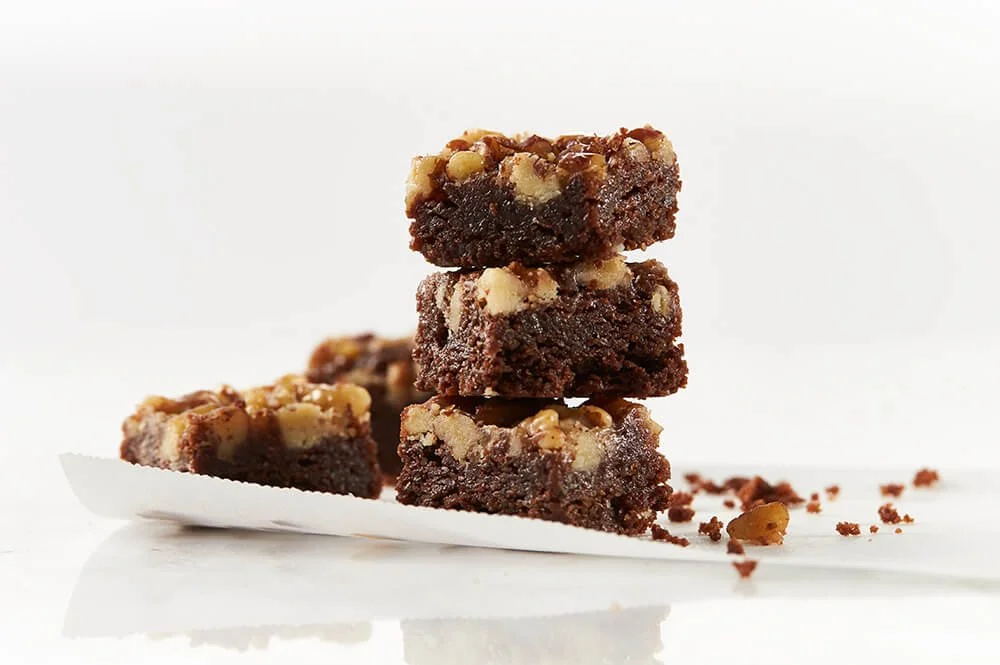 Stacked chocolate brownies with chopped nuts on a white surface.