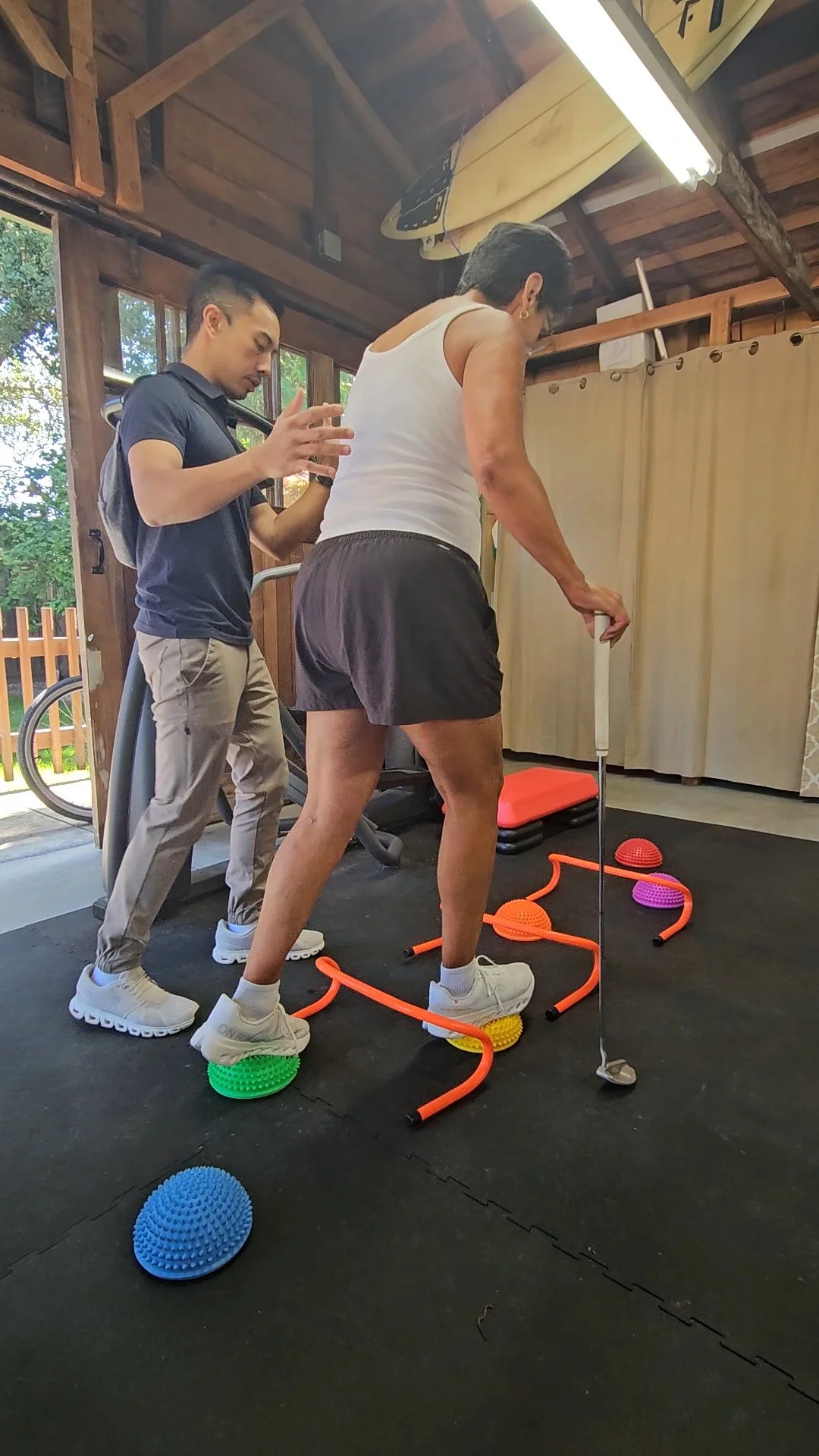 A woman practicing golf indoors on a workout mat with balancing discs, while a trainer observes and offers assistance. The room has wooden walls and natural light coming through the window.