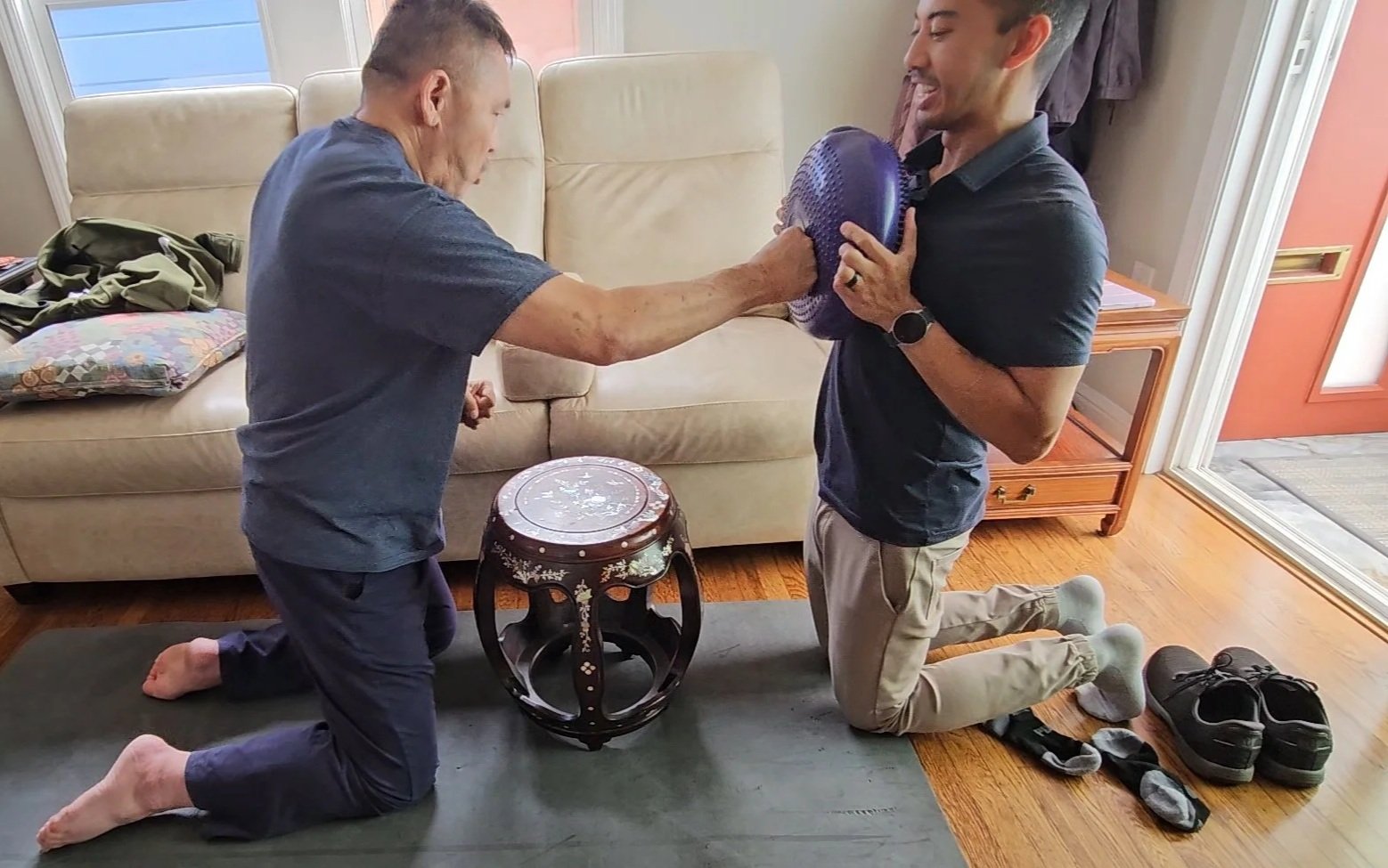 Two men kneeling on a rug in a living room, one older man with his eyes closed and an older woman's hand on his head, and a younger man holding a purple cardio ball close to his chest. The older man is reaching out to the younger man, who is smiling. Clothes, shoes, and a bag are on the floor and a small wooden table is between them.