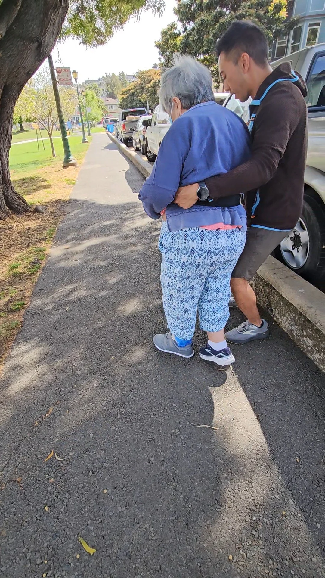 A young man helping an elderly woman walk on a sidewalk in a park-like area with parked cars and trees.