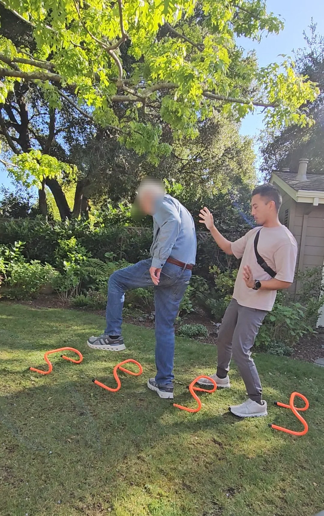 A young man is practicing a high kick in a backyard with two orange agility poles laid out on the grass. An older man stands nearby, watching and raising his hand to stop or guide him. The scene is set outdoors with greenery and trees in the background on a sunny day.