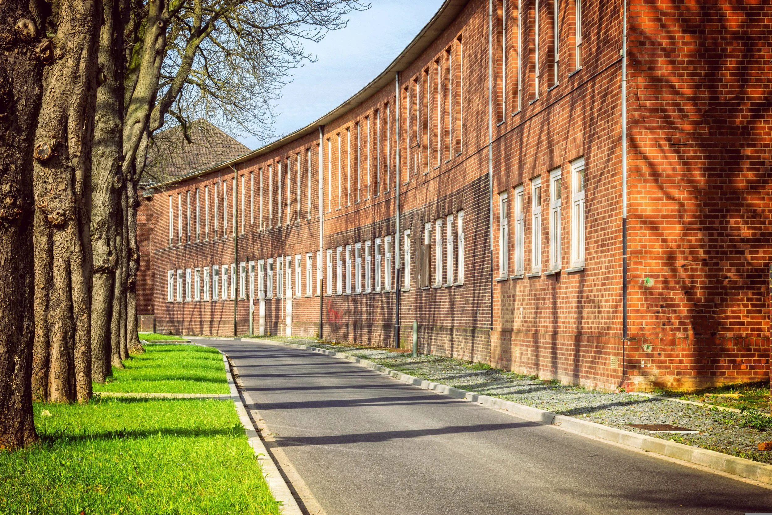 A long two-storey red brick building with rows of tall white windows curves alongside a quiet road, with trees and a strip of grass on the other side.