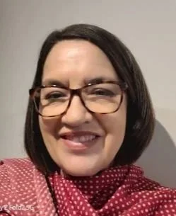 Close-up of a woman with shoulder-length dark hair, glasses, smiling, wearing a pink polka dot blouse.