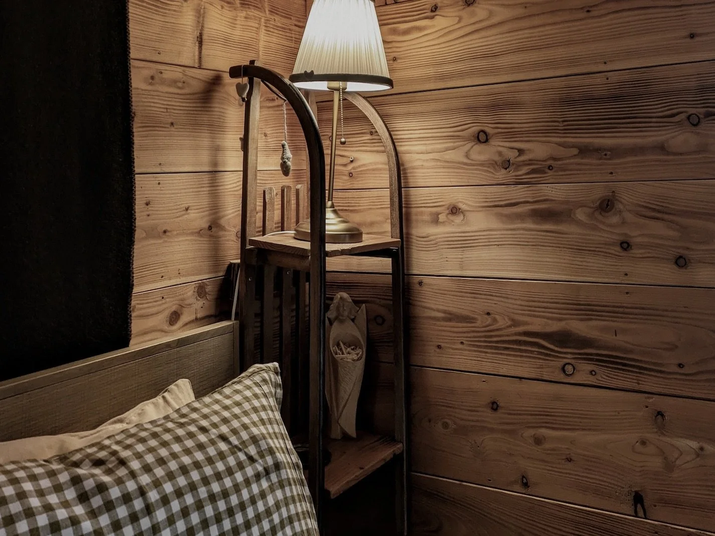A cozy bedroom corner with a bed, checked pillow, and a wooden wall. There is a nightstand with a lamp on top, and a decorative wooden item on the shelf.