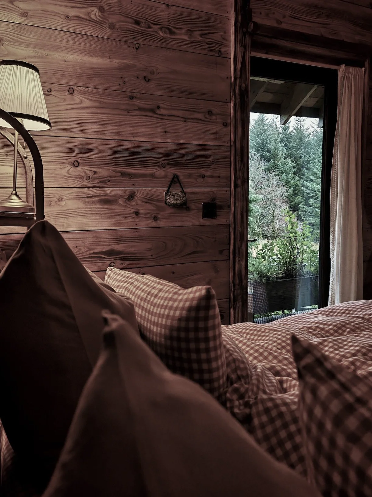 Cozy bedroom with wooden wall, bed with checkered and plain pillows, a lamp on the side, and a glass door leading to greenery outside.