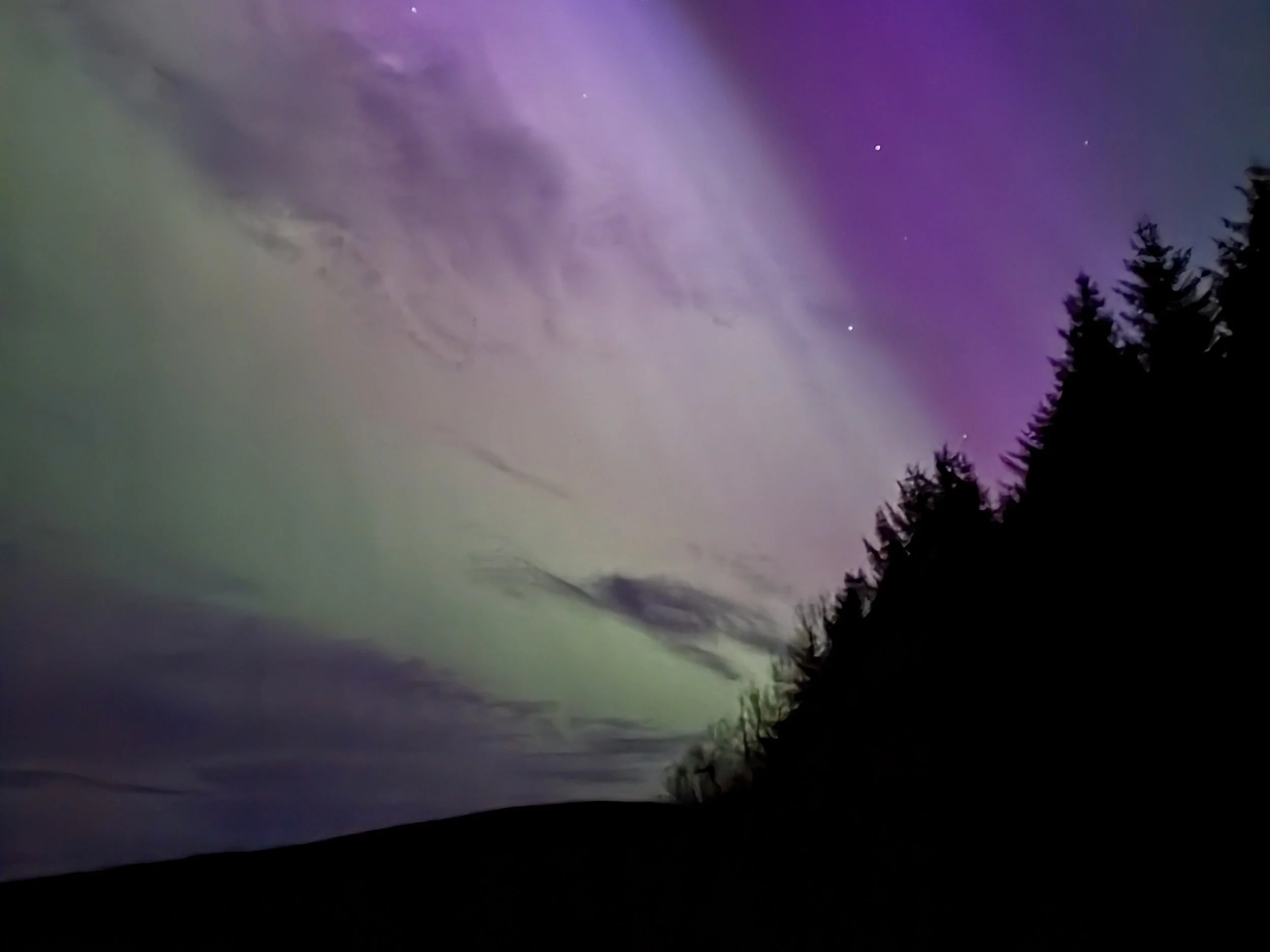 Night sky with aurora borealis, clouds, and visible stars over a silhouette of a forested hillside.