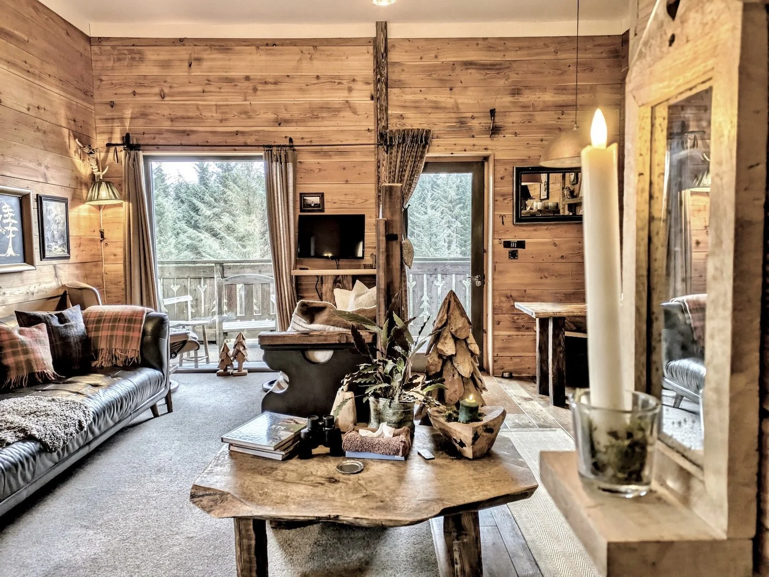 Cozy living room with wood-paneled walls, a leather sofa with plaid pillows, a wooden coffee table with decorative items, a wall-mounted television, large windows with curtains, and a balcony door.