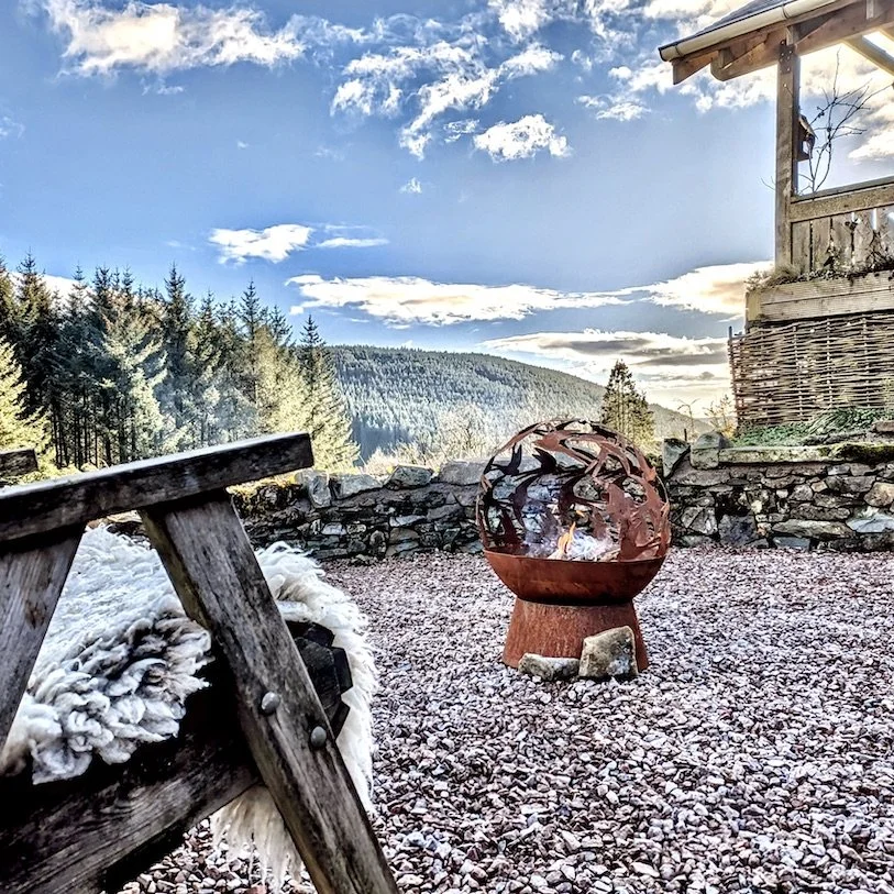 Outdoor scene featuring a rustic firepit on a gravel surface, a wooden fence, a mountain landscape with evergreen trees, and a partly cloudy sky.