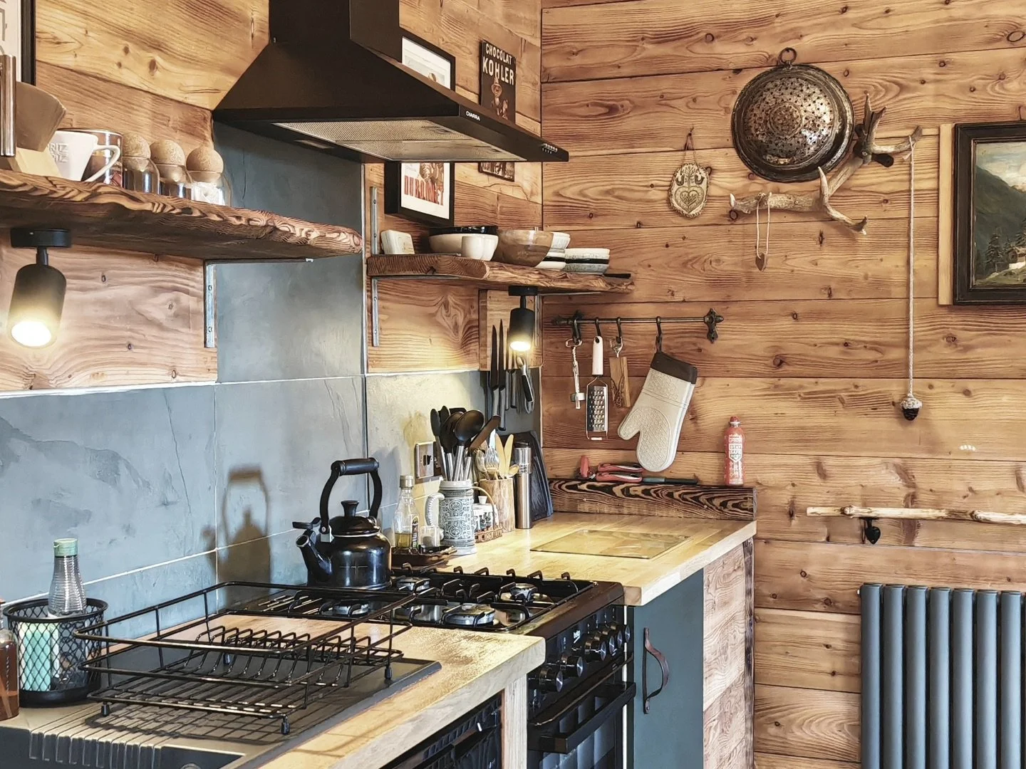 Cozy kitchen with wooden walls, open shelves holding bowls and jars, a black stove with kettle, knives, utensils, and a clock on the wall.