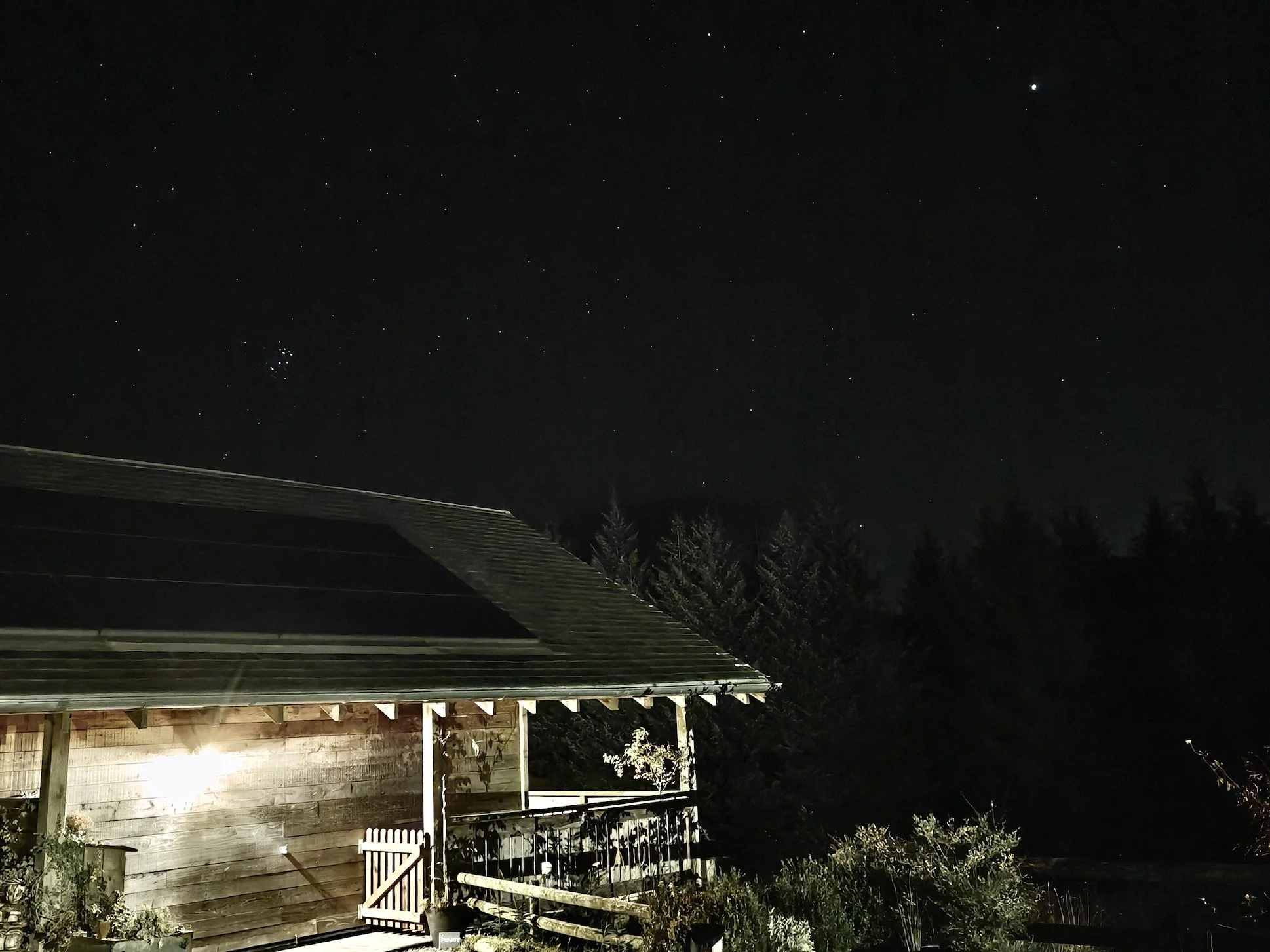 Nighttime scene of a wooden house with a porch and trees in the background under a starry sky.