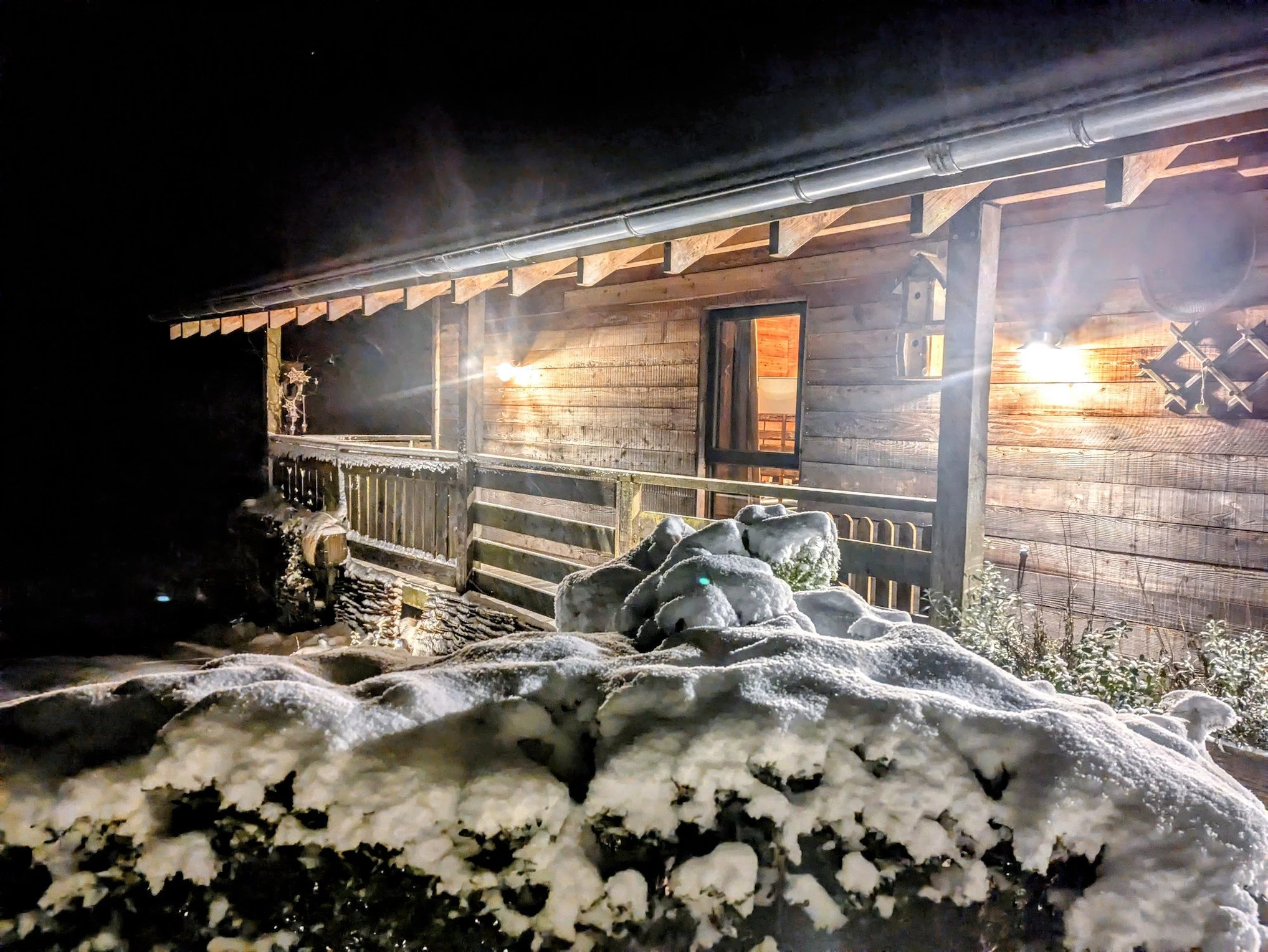 Nighttime view of a wooden house with a snow-covered porch, warm exterior lights, and snow piled in front.