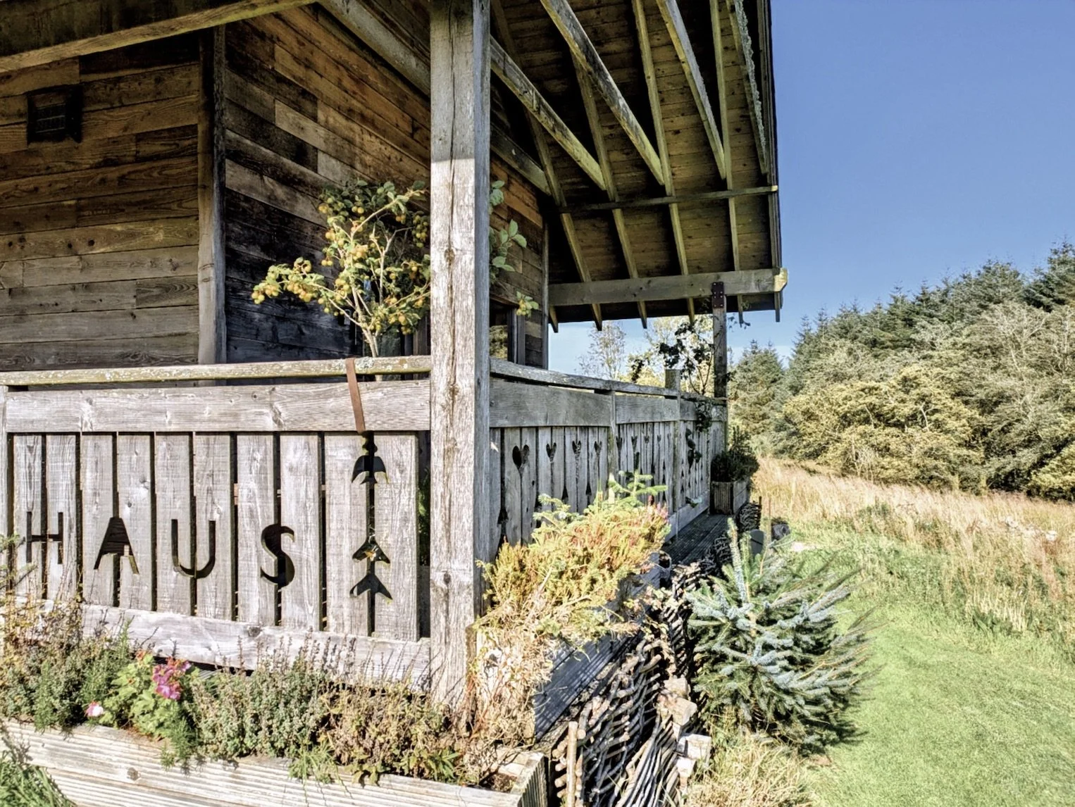 A rustic wooden farmhouse with a balcony decorated with plants and a metal sign spelling 'HYSU' hanging from the railing, set in a grassy countryside with trees in the background under a clear blue sky.