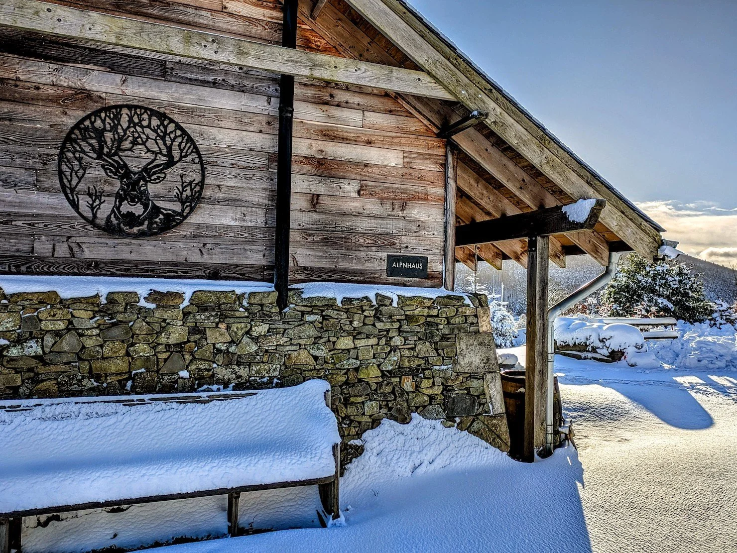 A rustic wooden building with a stone foundation covered in snow, featuring a black metal deer head and antlers sign on the side, a black downspout, and snow-covered ground and landscape in the background.