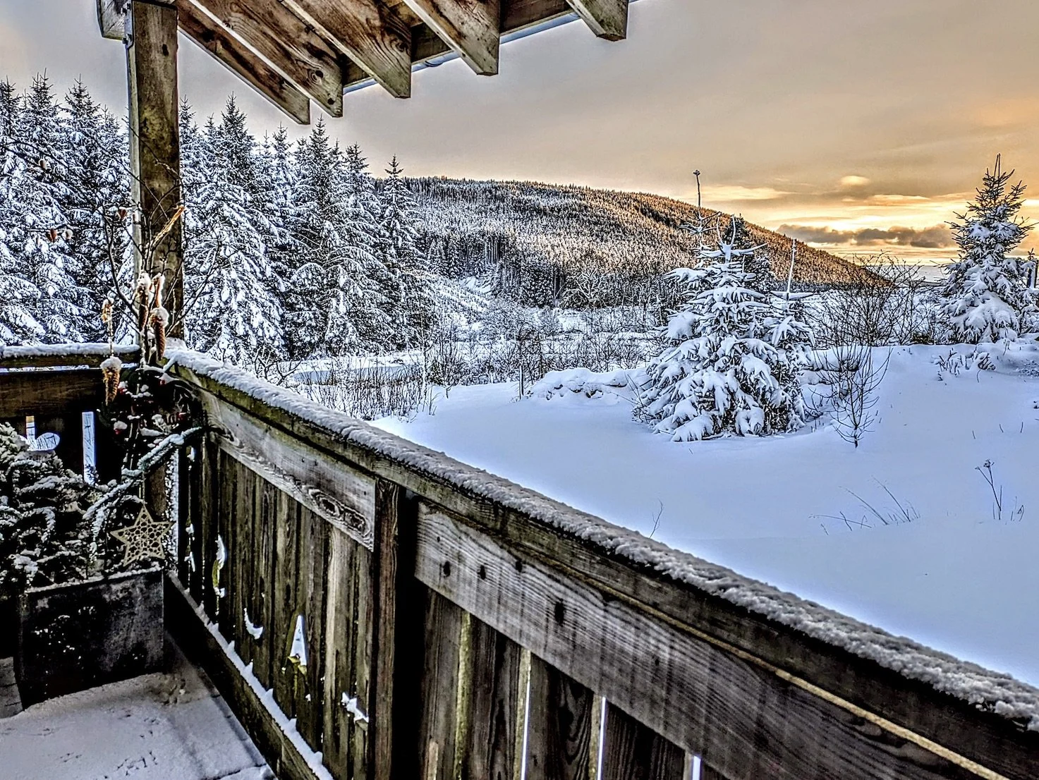 Snow-covered landscape viewed from a wooden balcony, with snow-laden trees and a mountain in the background under a partly cloudy sky at sunset.