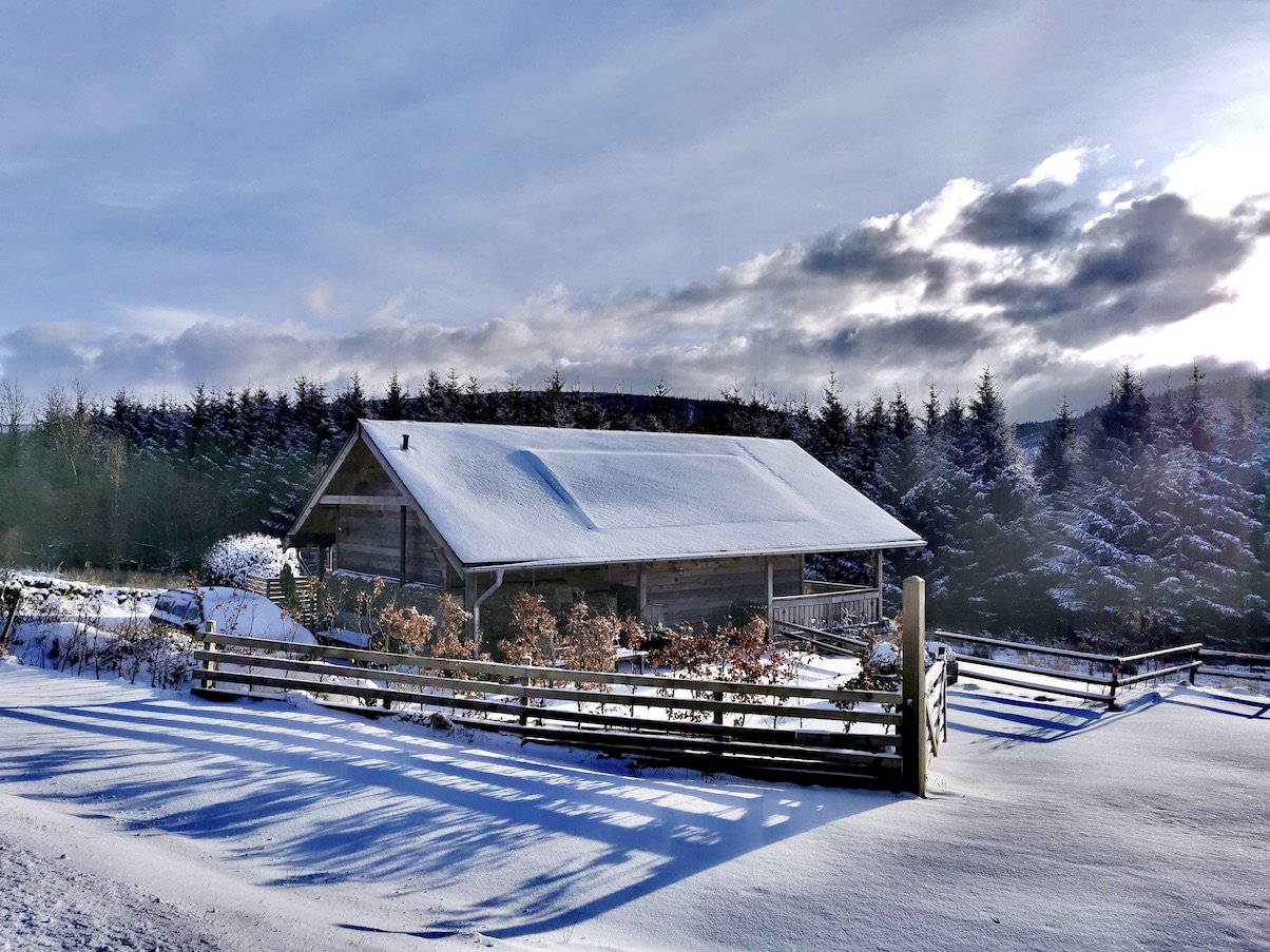 Snow-covered house in a winter landscape with a wooden fence, surrounded by snow-covered trees and a cloudy sky.