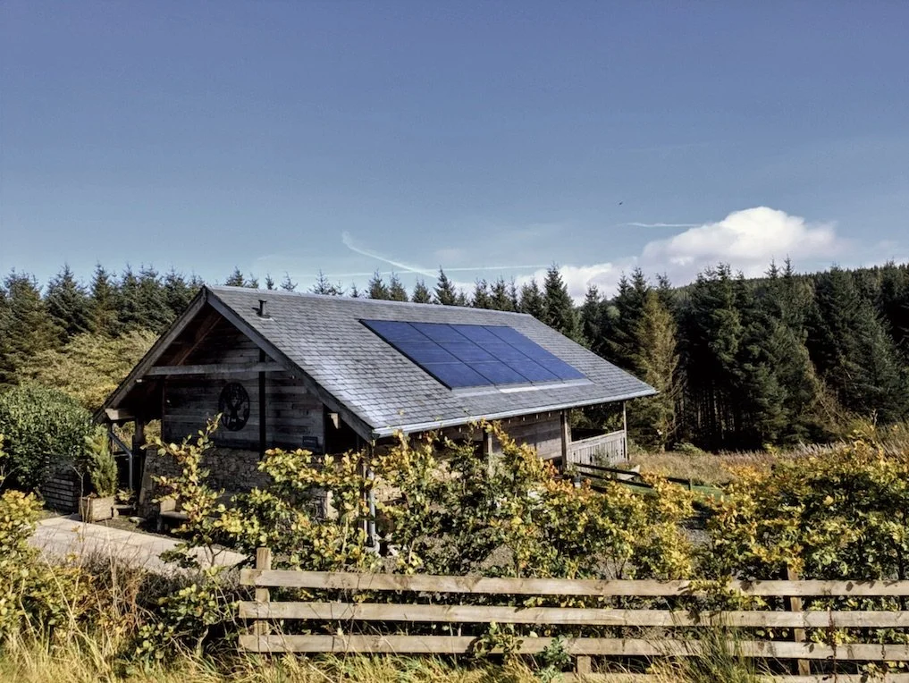 A rustic wooden house with solar panels on the roof, surrounded by greenery and a wooden fence, with trees and a blue sky in the background.