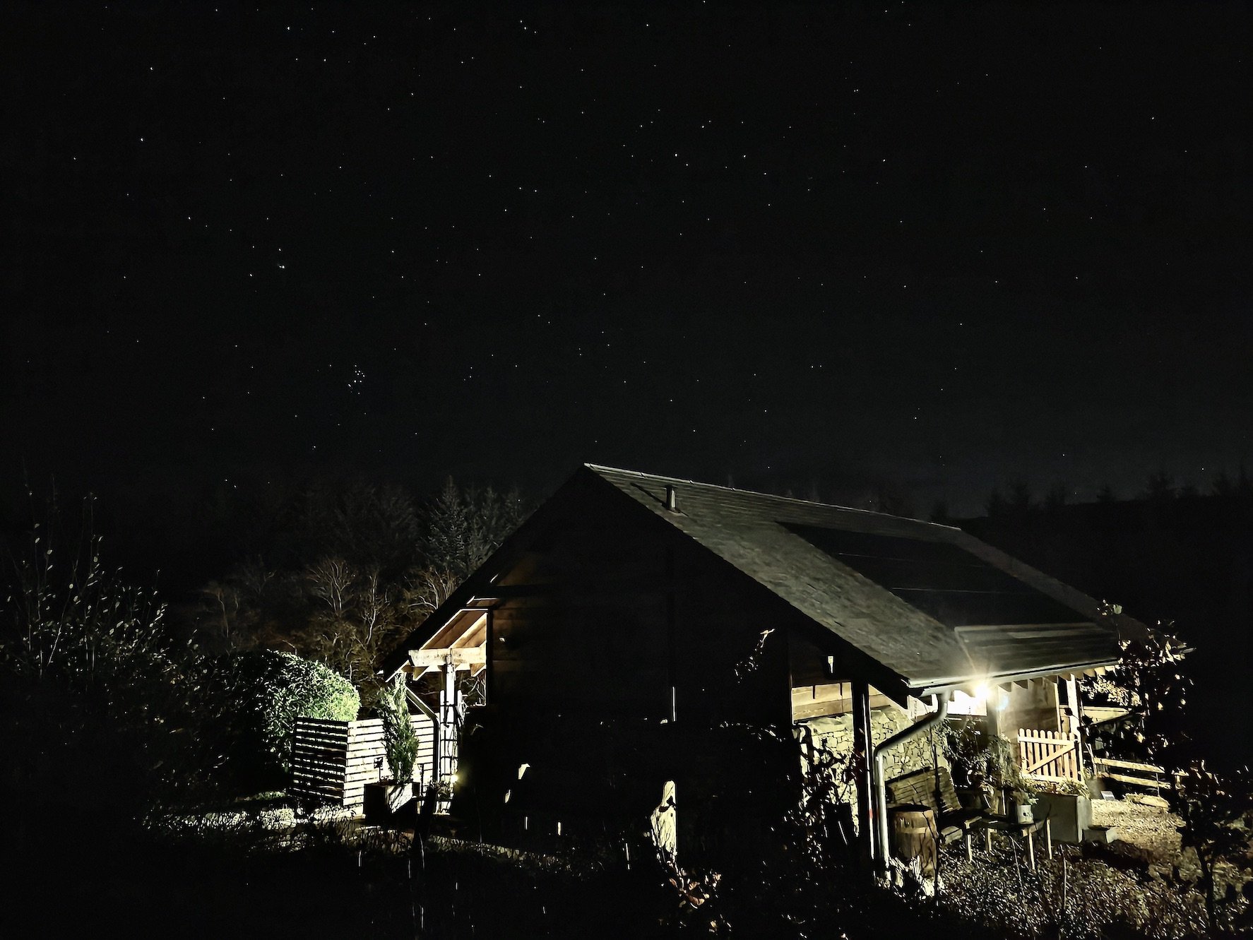 Nighttime scene of a small wooden house with a sloped roof illuminated from inside, surrounded by a garden with bushes and plants, and a starry sky overhead.