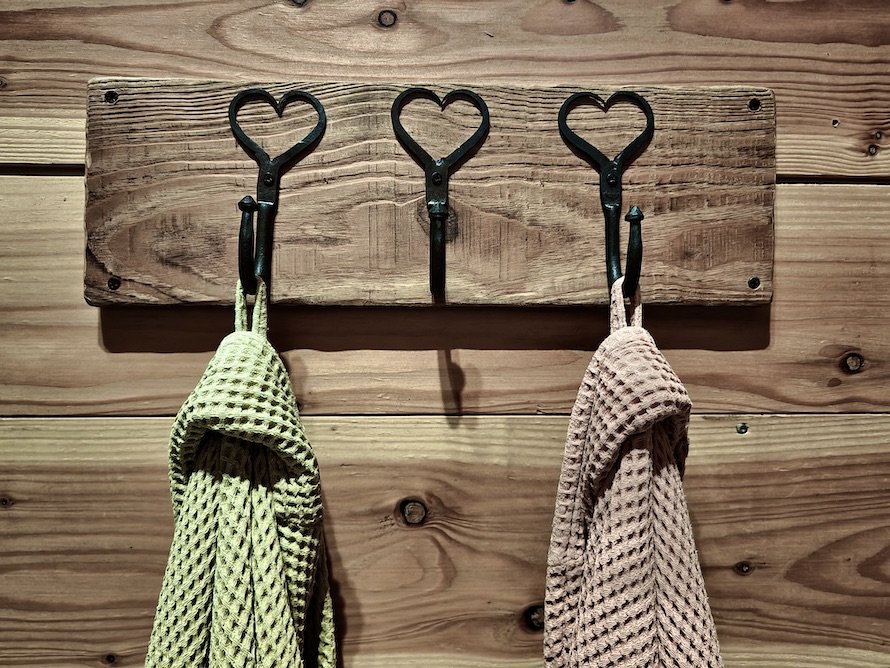 Wooden wall with a mounted wooden board holding three decorative metal hooks, each shaped like a heart, with kitchen towels hanging from two of the hooks.