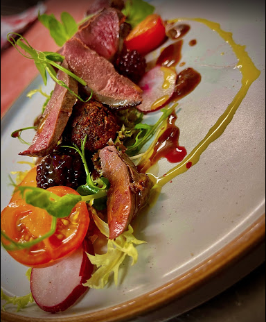 A plated dish featuring sliced medium-rare steak, cherry tomatoes, mixed greens, radish slices, blackberries, and drizzles of sauce and oil on a white oval plate.