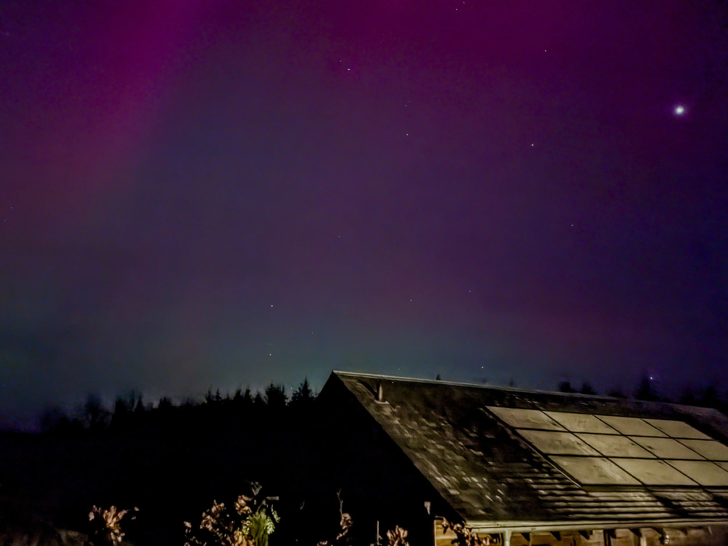 Night sky with stars, including a bright planet, above a house roof and silhouette of trees.