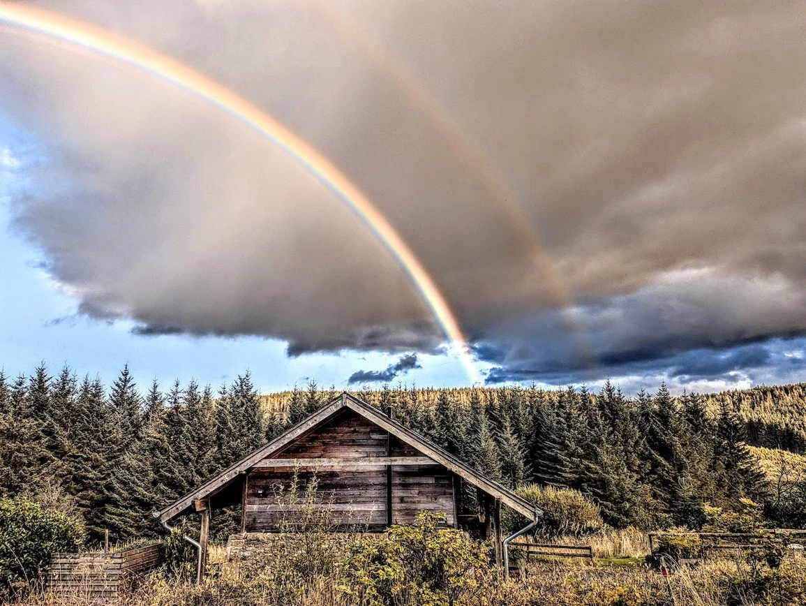 A double rainbow over a rustic wooden house surrounded by trees and hills.