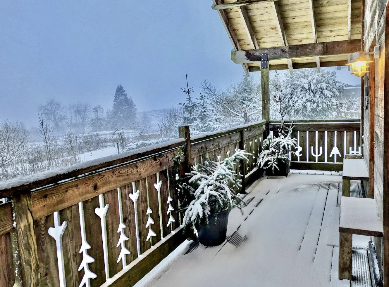 A snow-covered balcony with potted plants, a wooden railing with cut-out designs, and a snow-covered landscape with trees in the background.