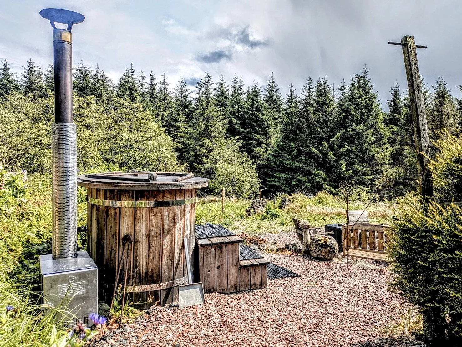 Wooden hot tub with stairs beside a gravel pathway, surrounded by lush greenery and trees under a cloudy sky.