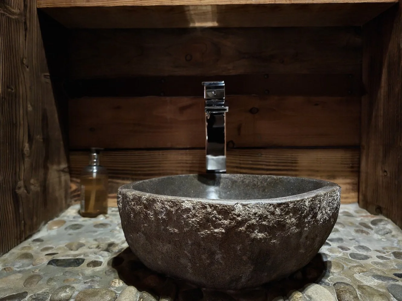 Stone bathroom sink with a modern chrome faucet, set against a wooden wall with a pebble stone mosaic countertop, and a soap dispenser to the left.