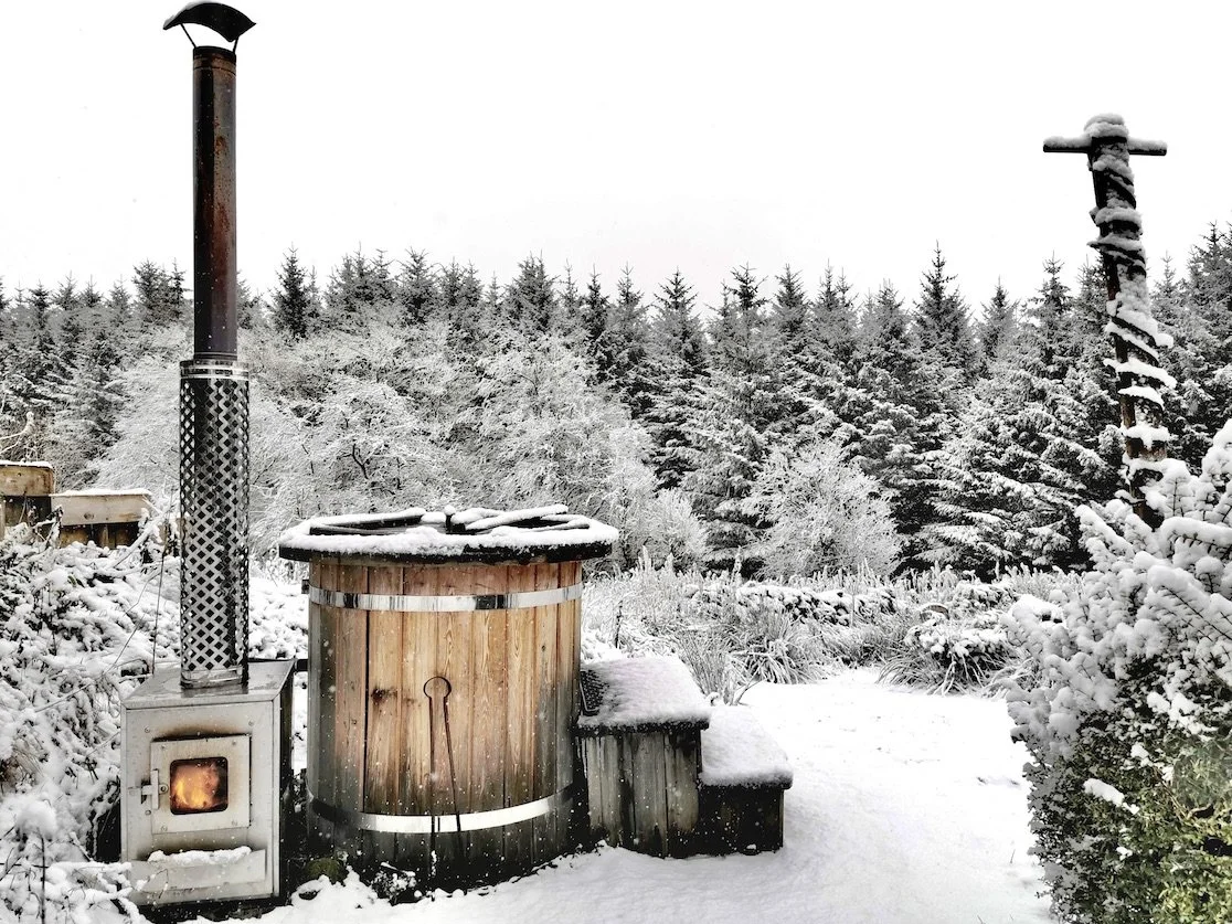 Snow-covered outdoor hot tub with a stove and chimney, surrounded by snow-covered trees in winter.
