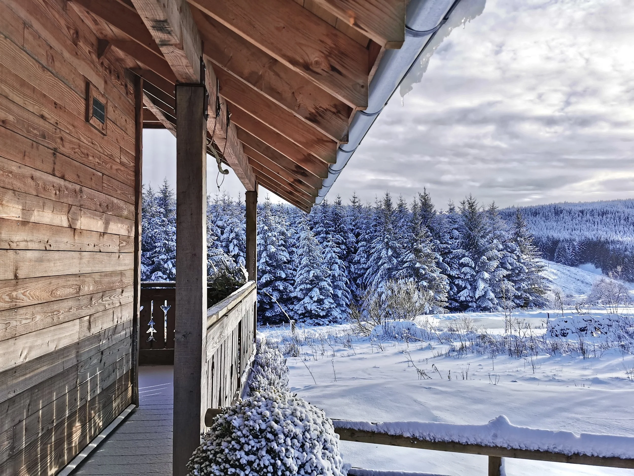 View from a wooden cabin porch overlooking a snow-covered landscape with trees and mountains in the distance, under a partly cloudy sky.