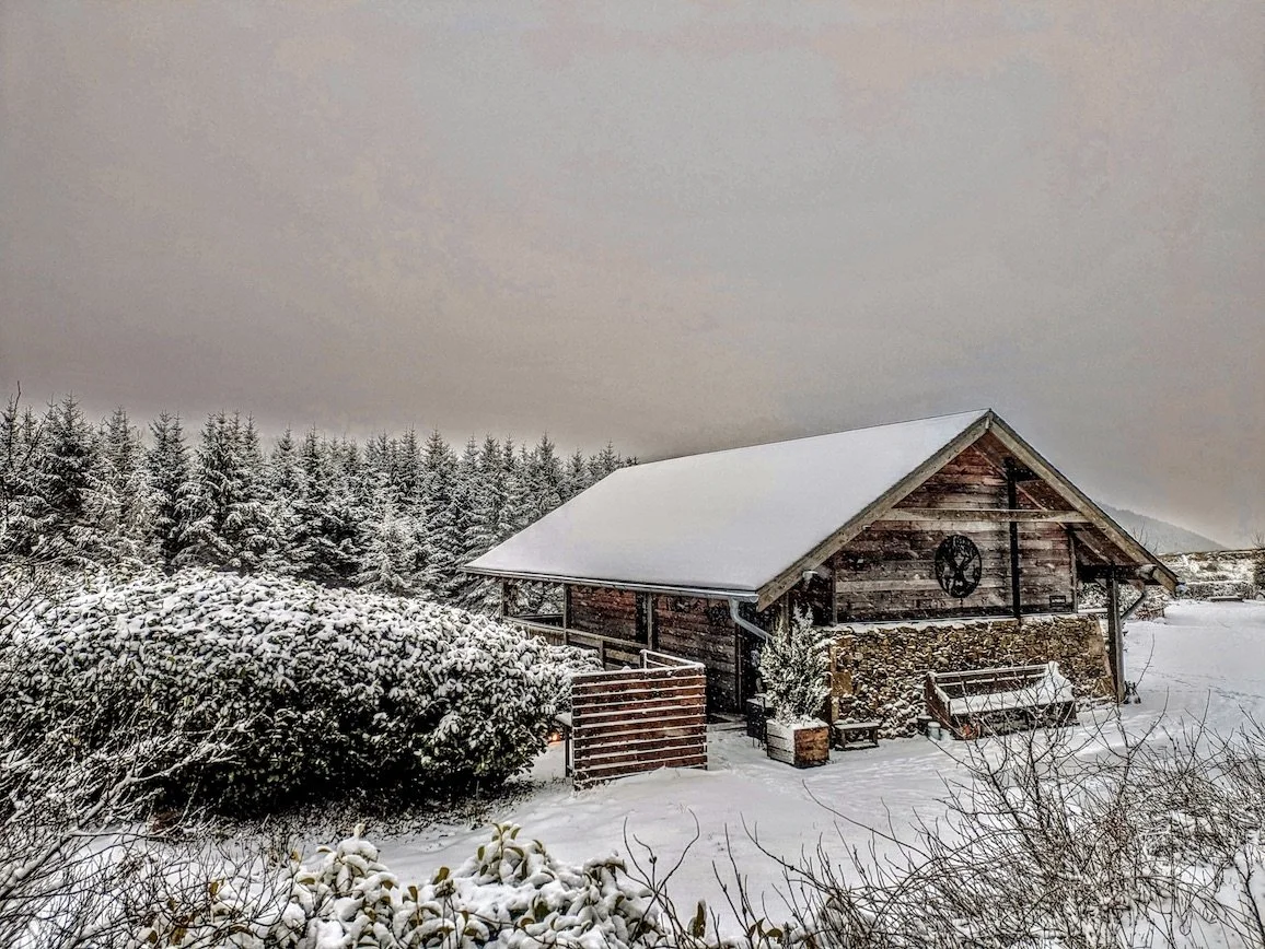 A rustic wooden barn with a snow-covered roof, surrounded by snow-covered trees and bushes in a winter landscape.