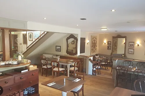Inside a cozy restaurant with wooden floors, elegant chairs, and framed artwork on the walls. There is a staircase leading upstairs, and a mirror with decorative framing. The space is warmly lit and inviting.
