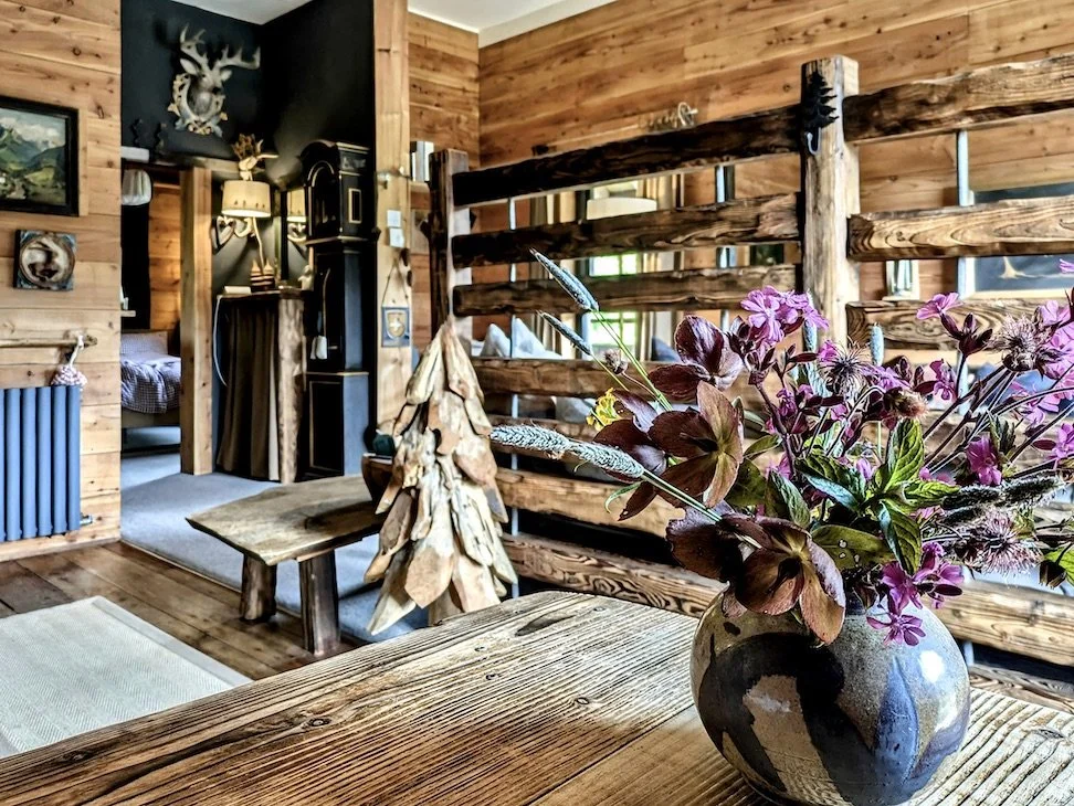 A rustic cabin interior with wooden walls, a vase of pink and purple flowers on a wooden table, and wooden furniture and decor.