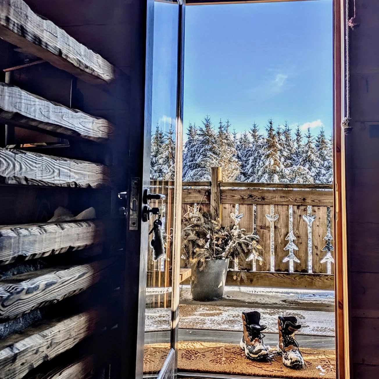 Open door to a snowy outdoor patio with a potted pine tree, wooden fence decorated with snowflake-shaped ornaments, and snowy mountain trees in the background. Snow boots are on a doormat inside.