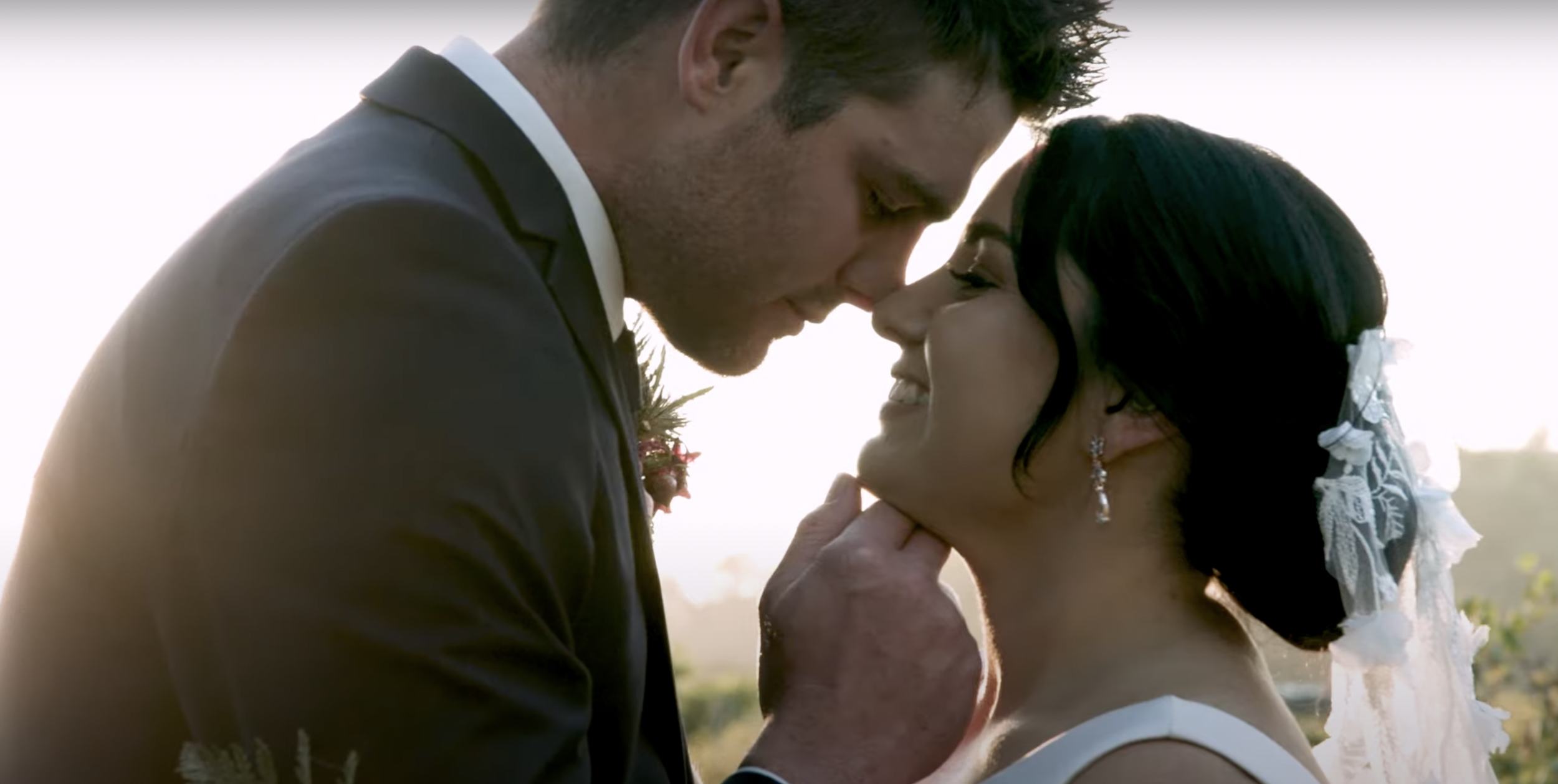 A couple in wedding attire sharing a close, intimate moment outdoors, with their foreheads and noses touching.
