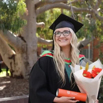 A young woman in a graduation cap and gown holding a bouquet of flowers and a diploma outside in a park.