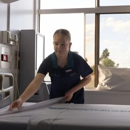 A young girl is making a bed with hospital sheets in a hospital room with large windows and sunlight outside.