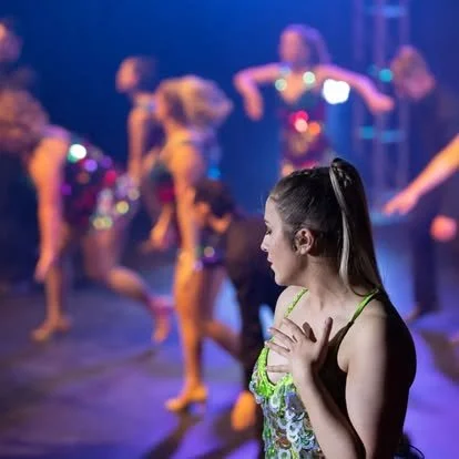 A young girl in a colorful dance costume stands in the foreground with other dancers blurred in the background during a dance rehearsal or performance.