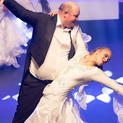 A man in a suit and a woman in a white dress dance together on stage, holding hands and moving gracefully.