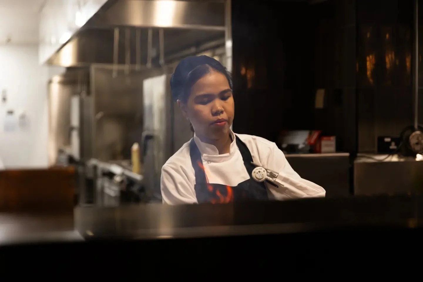 A female chef in a white jacket and black apron working in a professional kitchen with stainless steel appliances.