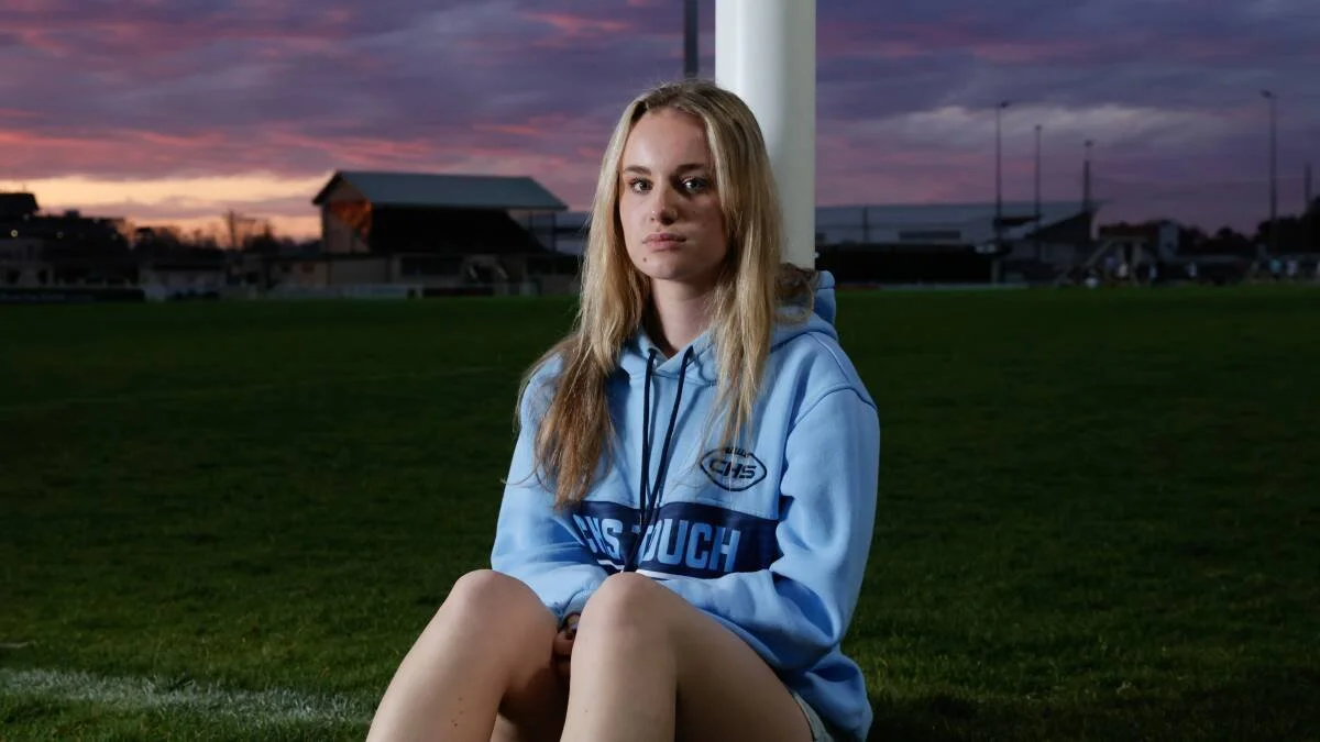Young woman sitting on a grassy sports field at sunset, wearing a light blue hoodie, with a serious expression.