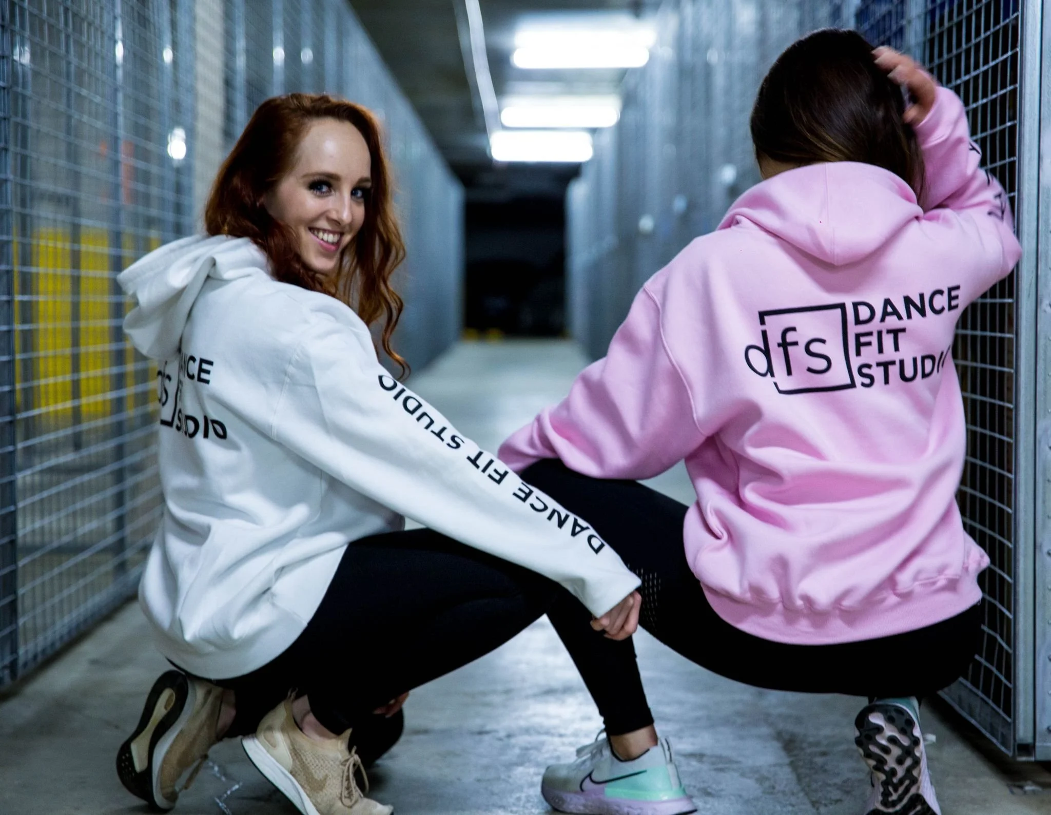 Two women are crouching in an indoor industrial-style hallway, wearing hoodies that say "Dance Fit Studio". One woman is smiling at the camera, wearing a white hoodie and beige sneakers, while the other woman, in a pink hoodie, has her head down and 