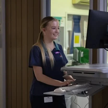 Young woman in a navy uniform smiling at a computer station in a medical facility.