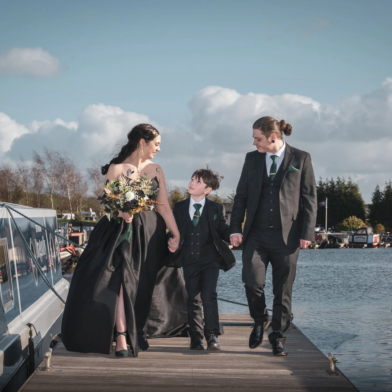 newly married couple walk with their son happily towards the camera during a couples photoshoot after their wedding at the boathouse at aston marina in stone staffordshire
